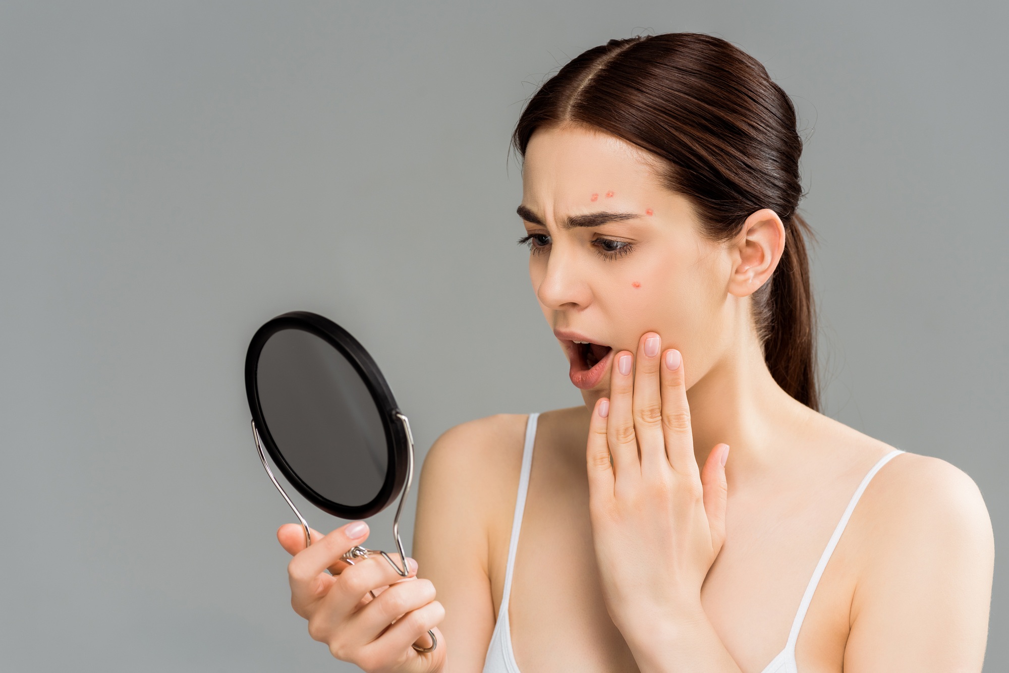 shocked young woman with acne on face looking at mirror isolated on grey