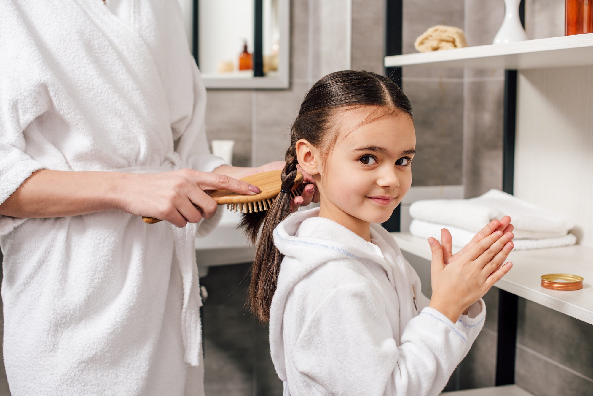 mother in white bathrobe combing daughter hair with wooden hairbrush near shelves in bathroom