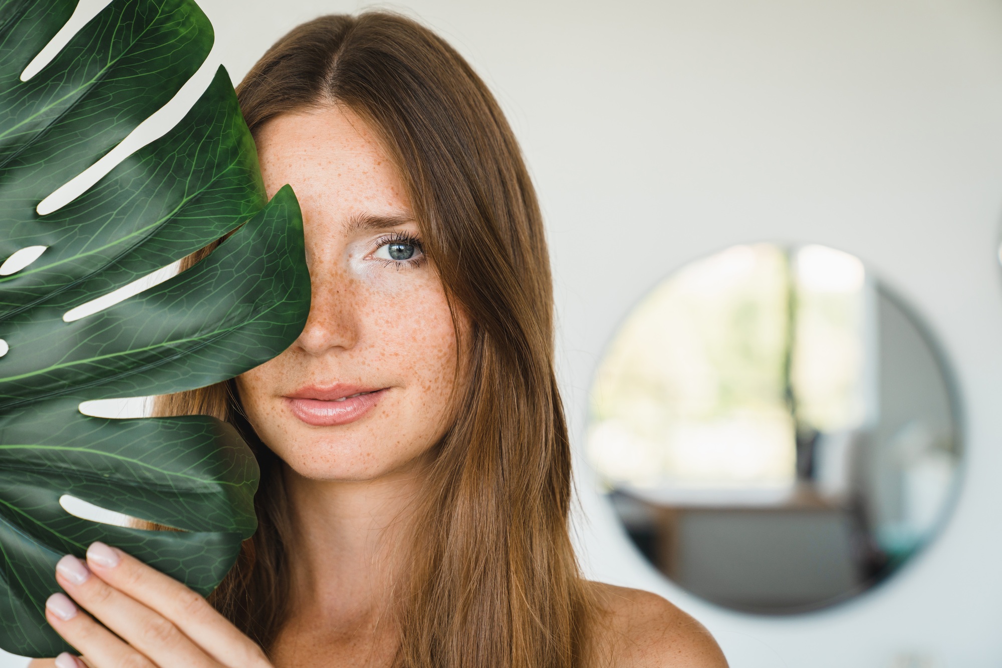 Young beautiful woman covering her face with green leaf at home. Beauty and skin care concept