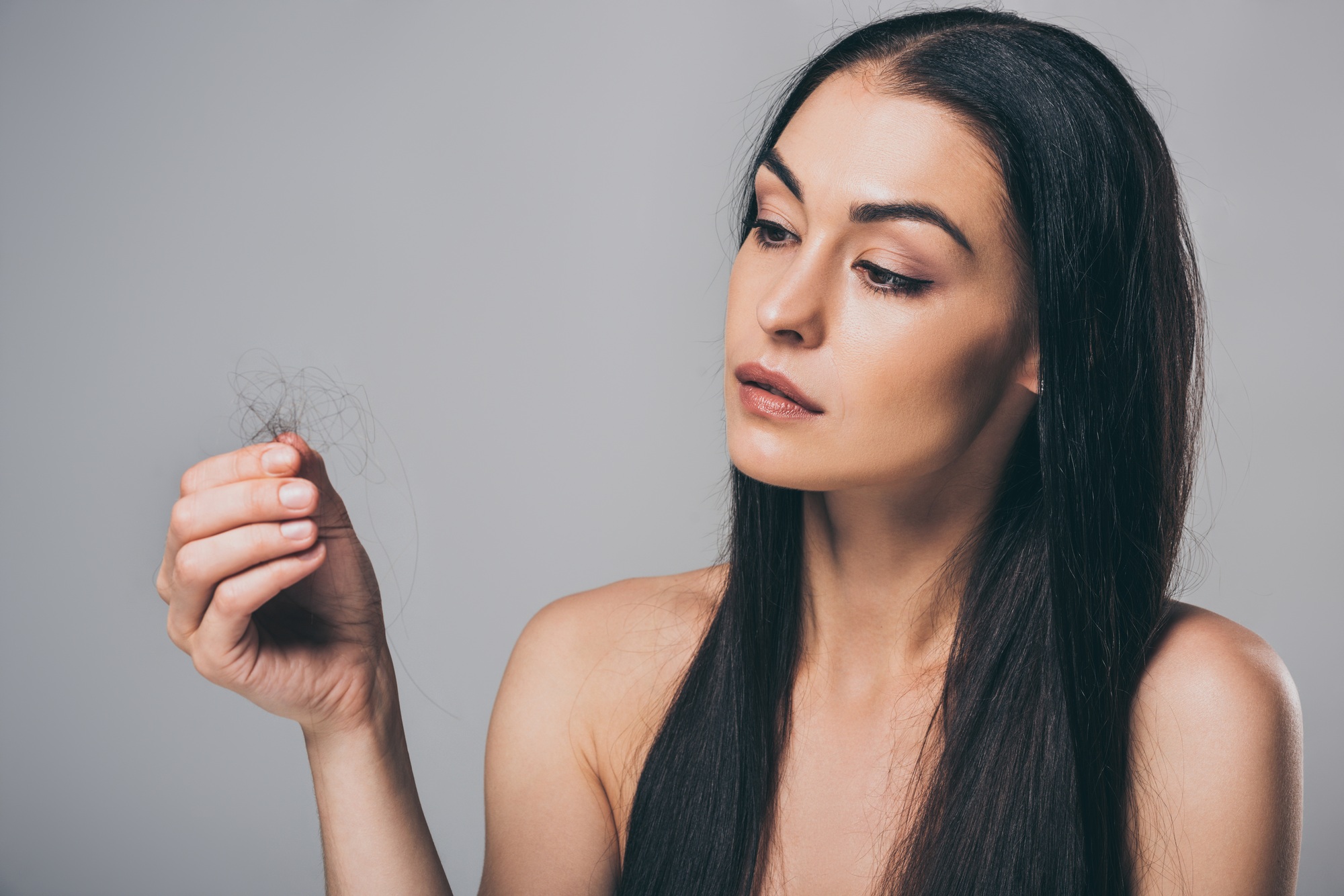 young brunette woman holding fallen hair isolated on grey, hair loss concept