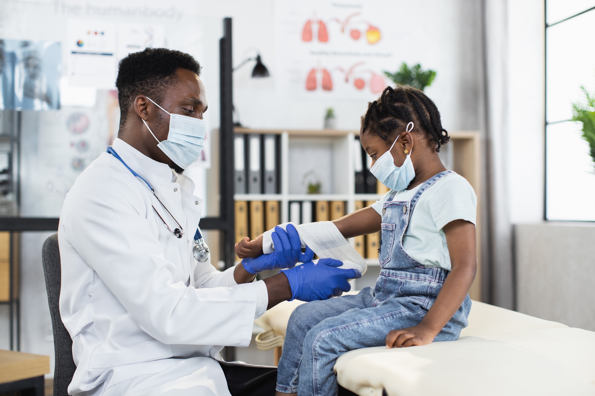 African doctor bandaging arm of little mixed race girl at modern clinic