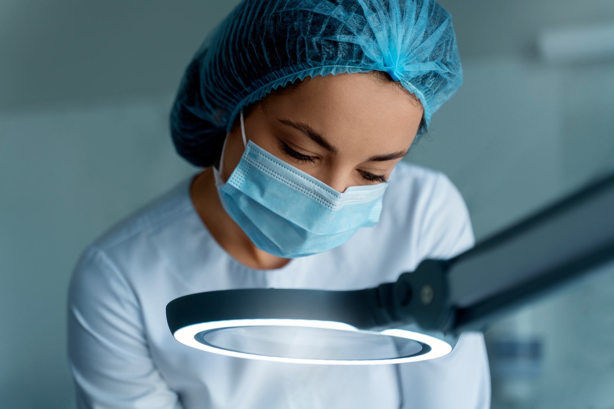 Cropped female cosmetologist looking at client's face through magnifying lamp