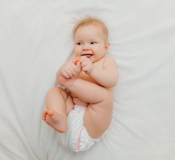 a happy joyful baby in diapers is lying on a white bed and holding his leg. High-quality photo.